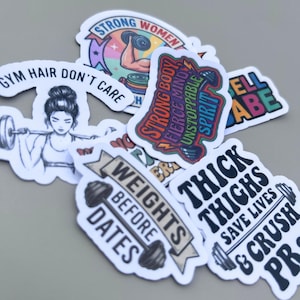Various powerlifting decals for women scattered across a neutral gray surface. The vinyl decal set includes diverse designs with motivational text and fitness graphics, perfect for a funny gym decal collection used to personalize water bottles or laptops with a unique aesthetic.