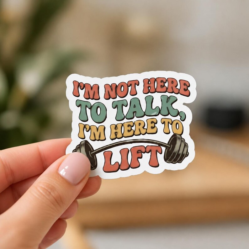 A hand with light pink nail polish holding a white sticker with a wavy border and the text I'M NOT HERE. This close-up shows the unique shape and humorous text of the funny gym decal included in the 9 pack.