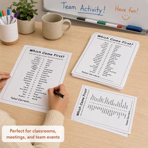 A top-down view of a wooden desk featuring several printed 'Which Came First?' trivia game sheets, a person's hands holding a pen over one sheet, a white mug, a small potted plant, and a whiteboard in the background. This Easy Party Starter Game works great for office gatherings.