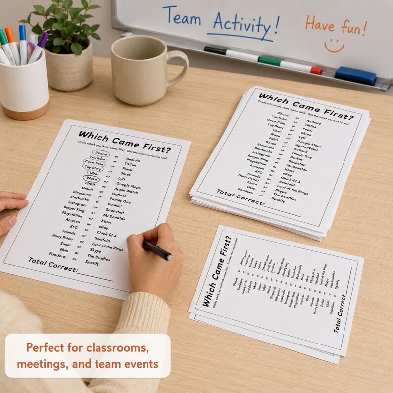 A top-down view of a wooden desk featuring several printed 'Which Came First?' trivia game sheets, a person's hands holding a pen over one sheet, a white mug, a small potted plant, and a whiteboard in the background. This Easy Party Starter Game works great for office gatherings.