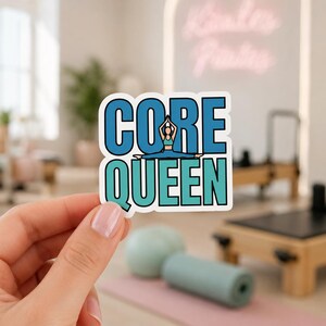 A hand with light pink manicured nails holding a sticker that says CORE QUEEN in large stylized letters. This die cut vinyl sticker is a great way to show off core strength and motivation on your gym locker or water bottle.