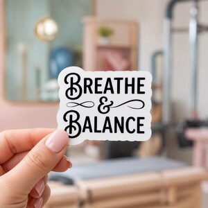 A hand holding a white sticker with the decorative script text BREATHE & BALANCE. This calming design is a beautiful part of our pilates sticker pack, ideal for anyone seeking peace and balance through their fitness practice.