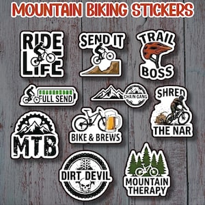 May include: A collection of mountain biking stickers with various designs and text. The stickers feature images of mountain bikers, mountain bike graphics, and phrases like "Ride Life", "Send It", and "Trail Boss". The stickers are on a wooden background.