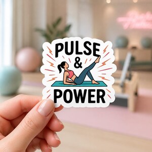 A hand holding a sticker that says PULSE & POWER with an illustration of a woman in pink workout gear. This dynamic sticker is a great way to celebrate movement and is perfect for decorating a cell phone case or journal.