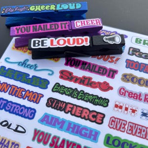 Close-up product photo of decorated cheer clothespins on top of a colorful cheer sticker sheet. Shows finished clips with sayings like “Fly High Cheer Loud,” “You Nailed It,” and “Be Loud,” along with the sticker sheet underneath for DIY cheer clip decorating and team spirit ideas.