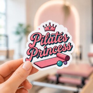 A hand holding a pink and teal sticker that says Pilates Princess in a beautiful cursive font. This close-up shows the vibrant colors of the pilates princess stickers, perfect for use as a cell phone case decoration or planner sticker.