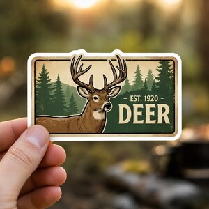 A hand holds a vintage deer sticker with a white border, featuring a classic deer illustration against a green forest background. The sticker includes the text EST. 1920 and DEER. This close-up shows the detail of the rustic hunting stickers included in the vintage wildlife sticker pack, perfect for laptop decals or water bottles.