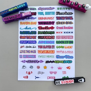 Cheer clip sticker sheet displayed with decorated wooden clothespins using phrases like “Cheer Loud,” “You Nailed It,” “Hit Zero,” and “Be Loud.” A bright cheer-themed product photo showing how the stickers fit on clips for team spirit pins, cheer bags, and competition favors.