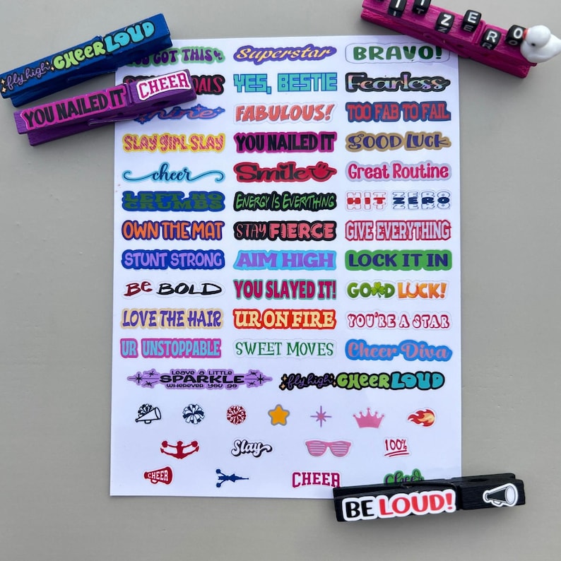 Cheer clip sticker sheet displayed with decorated wooden clothespins using phrases like “Cheer Loud,” “You Nailed It,” “Hit Zero,” and “Be Loud.” A bright cheer-themed product photo showing how the stickers fit on clips for team spirit pins, cheer bags, and competition favors.