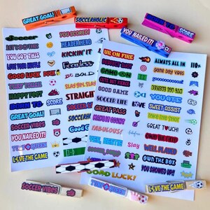 Two white sheets of colorful soccer-themed stickers and several decorated clothespins in bright blue, pink, and orange featuring phrases like Great Goal, Soccerholic, and Born to Score. These clothes pin youth accessories are a hit at sports camps and tournaments. They allow young athletes to showcase their personality while supporting their teammates on the field. These vibrant designs add a pop of color to any gear.
