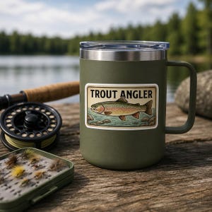 A Trout Angler sticker from the fishing decal set is displayed on an olive green mug in a rustic fishing scene. The mug sits on a wooden dock near a rod, reel, and fly box overlooking a lake. This mockup demonstrates how the vintage wildlife sticker pack can enhance outdoor gear like mugs and tackle boxes for an angler.