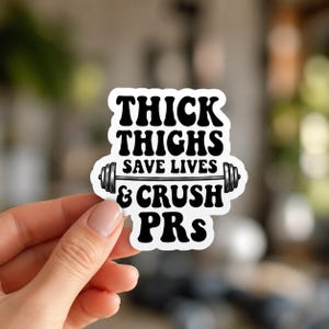 A hand holding a white sticker with black text and a barbell graphic that reads THICK THIGHS SAVE LIVES & CRUSH. This humorous design is a standout piece in the powerlifting decals for women collection.