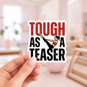 A hand holding a white sticker with the bold text TOUGH AS A TEASER. This high-quality die cut vinyl sticker is designed for those who embrace the challenge of mat pilates and want to display their strength on their gear.