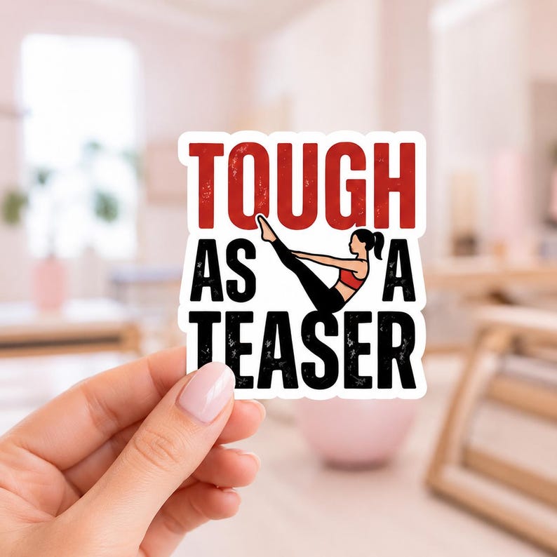 A hand holding a white sticker with the bold text TOUGH AS A TEASER. This high-quality die cut vinyl sticker is designed for those who embrace the challenge of mat pilates and want to display their strength on their gear.