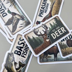 Rustic hunting stickers displayed in a close-up, top-down view on a light gray surface. This fishing decal set includes die-cut vintage-style wildlife stickers such as a deer, bass, pheasant, and duck. The stickers feature classic text like DEER EST. 1920 and BASS FISHING, making them a perfect vinyl decal set for planners, laptops, or water bottles for any nature lover.