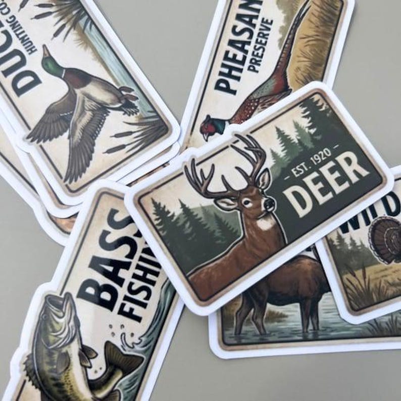 Rustic hunting stickers displayed in a close-up, top-down view on a light gray surface. This fishing decal set includes die-cut vintage-style wildlife stickers such as a deer, bass, pheasant, and duck. The stickers feature classic text like DEER EST. 1920 and BASS FISHING, making them a perfect vinyl decal set for planners, laptops, or water bottles for any nature lover.
