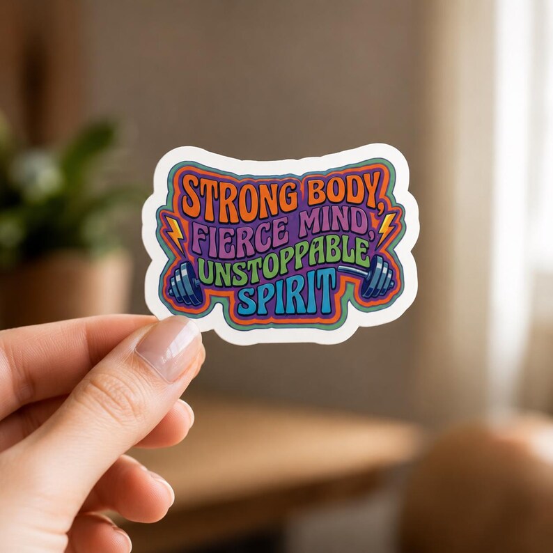 A hand holding a colorful sticker with the text STRONG BODY, FIERCE MIND, UNSTOPPABLE SPIRIT in a stylized font. This close-up demonstrates the vibrant colors and motivational message of the fitness gym sticker set.