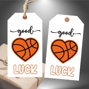 May include: Two white gift tags with a basketball heart design. The word "good" is written in a cursive font above the heart, and the word "LUCK" is below it in orange block letters. The tags are attached to a gift.