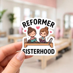 A hand holding a white sticker featuring a cartoon illustration of two girls sitting on a Pilates reformer machine. This funny reformer stickers design is a charming addition to any pilates princess kit or fitness enthusiast's collection.