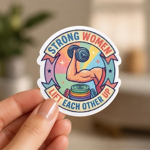 A hand holding a circular sticker featuring a muscular arm lifting a dumbbell with weight plates below. This close-up shot highlights the detailed graphic design of the female weightlifting stickers and their high-quality print.