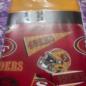 May include: A fabric sheet featuring a repeating pattern of San Francisco 49ers team logos, including football helmets, pennants, and the text 49ERS in red, gold, and white. The fabric has a solid gold band at the top and is encased in clear plastic.