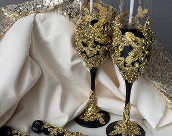 May include: Elegant champagne flutes and cake serving set. The flutes have black stems and are adorned with gold embellishments. The cake server set features black handles with matching gold accents. The background includes a gold sequined fabric.
