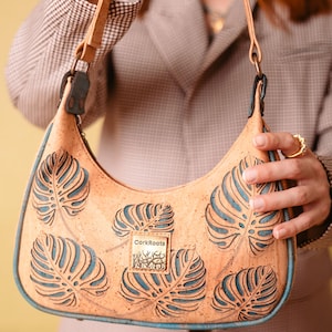 May include: A crescent-shaped handbag crafted from light tan cork material, featuring a cutout monstera leaf pattern that reveals a teal blue interior layer. A square metal plaque with the text CorkRoots is centred on the front surface.