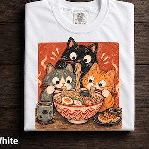 Peut inclure: Un t-shirt blanc avec une illustration graphique de trois chats mangeant des ramen dans un grand bol. Les chats sont noirs, gris et orange, utilisant des baguettes pour manger des nouilles. Le motif inclut une tasse, des raviolis et un fond rouge.