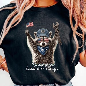 Könnte beinhalten: Ein schwarzes T-Shirt mit einer Grafik eines Waschbären, der eine blaue Mütze, eine Sonnenbrille mit US-Flaggenmuster und ein blaues Halstuch trägt. Der Waschbär hält eine kleine US-Flagge und einen Schraubenschlüssel. Text Happy Labor Day.