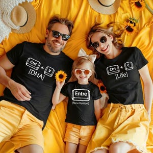 May include: A family wearing matching black t-shirts with white text. The shirts have keyboard-themed designs with the words "Ctrl + V [DNA]", "Entre [New Version]", and "Ctrl + C [Life]" printed on them. They are lying on a yellow blanket.