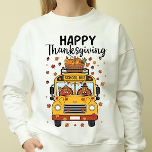 Op de afbeelding: Witte sweatshirt met een Thanksgiving-thema. Het ontwerp toont een gele schoolbus met twee kalkoenen als chauffeurs, een mand met pompoenen en kalebassen en herfstbladeren. De tekst "HAPPY Thanksgiving" is in het zwart gedrukt.