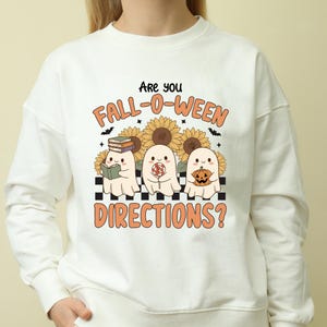 Puede incluir: Sudadera blanca con un gráfico que presenta tres fantasmas de dibujos animados frente a girasoles. El texto dice: "Are you Fall-O-Ween Directions?" Un fantasma sostiene un libro, otro una piruleta y el tercero una calabaza.
