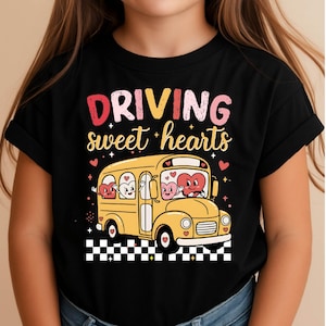 May include: Black t-shirt featuring the text "DRIVING sweet hearts" in pink, yellow, and red. A yellow school bus with heart-shaped passengers is the central graphic. The bus is set against a black and white checkered pattern.