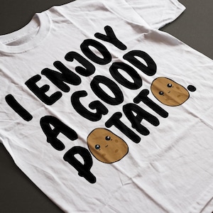 May include: White t-shirt with the phrase "I ENJOY A GOOD POTATO" in large, black, bold letters. Two cartoon potatoes with smiling faces are also featured. The shirt is made of a soft material.