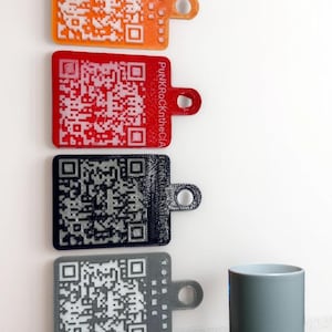 Puede incluir: Cuatro llaveros cuadrados con códigos QR impresos en diferentes colores. Los llaveros son naranja, rojo, negro y gris. Los llaveros están colgados en una pared blanca. Una taza cilíndrica gris está sobre una superficie blanca.