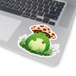 May include: A sticker featuring a cartoon frog wearing a mushroom hat. The frog is green with a light green belly and pink cheeks. The mushroom hat is tan with brown spots. The frog is sitting in green grass.