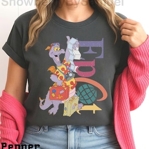 May include: Dark gray t-shirt featuring a purple dragon character with luggage, a camera, and the word "Figment" in colorful letters. The design includes a globe and an arrow, suggesting travel or adventure. The shirt is a casual wear item.