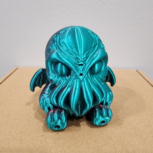 May include: A teal green 3D printed figurine of a Cthulhu-like creature with tentacles and wings. The figurine has a detailed, textured surface and is sitting on a brown surface.