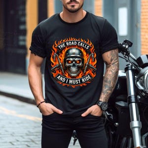 K&ouml;nnte beinhalten: Schwarzes T-Shirt mit einem Sch&auml;del-Design, der einen Helm tr&auml;gt, mit flammenden Augen und von Flammen umgeben ist. Der Text "THE ROAD CALLS AND I MUST RIDE" ist oberhalb und unterhalb des Sch&auml;dels zu sehen. Ein Motorrad ist teilweise rechts zu sehen.