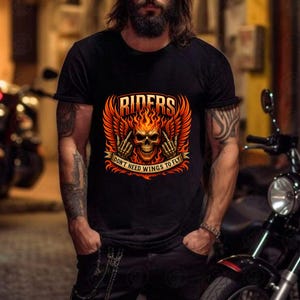 K&ouml;nnte beinhalten: Schwarzes T-Shirt mit einem flammenden Totenkopf-Design, dem Wort "RIDERS" dar&uuml;ber und dem Text "DON'T NEED WINGS TO FLY" darunter. Das Design beinhaltet orangefarbene und rote Fl&uuml;gel und Flammen. Das T-Shirt wird von einer Person getragen.