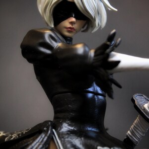 May include: A detailed figurine of a character with short white hair, a black blindfold, and a black outfit. The figure is posed with one arm extended, holding a sword. The outfit has intricate white floral patterns.