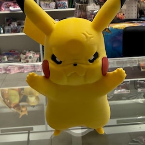 May include: A yellow Pikachu figurine with black-tipped ears, red cheek circles, and an angry expression. The figure has outstretched arms and is standing on a reflective surface. The background shows shelves with various items.
