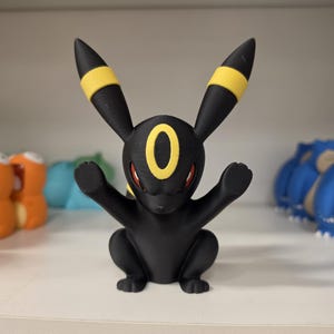 May include: A matte black figurine of the Pokemon character Umbreon, featuring long ears with yellow bands, a yellow oval marking on the forehead, and red eyes. The creature is depicted in a seated pose with both front paws raised upward.
