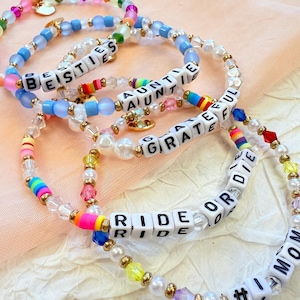 May include: Close-up of several beaded bracelets with white lettered cubes spelling out words like "BESTIES", "AUNTIE", "GRATEFUL", and "RIDE OR DIE". The bracelets feature colorful beads, pearls, and gold accents, creating a vibrant and personalized accessory.