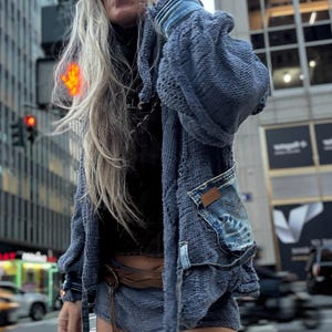 May include: A person wearing a blue knit hooded cardigan with denim pocket details, denim shorts, and sunglasses. The person is standing in a city street with buildings and a crosswalk in the background.