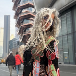 May include: A black crochet cardigan with colorful floral designs in red, green, blue, and yellow. The open-front cardigan is worn over a black top and blue jeans. The background features a modern architectural structure and pedestrians.