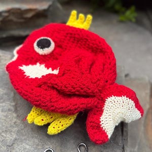 May include: A handmade red crocheted fish-shaped item with white and yellow accents. The fish has a black and white eye and a yellow crown. Two silver-coloured metal clasps with a chain are visible below the fish.