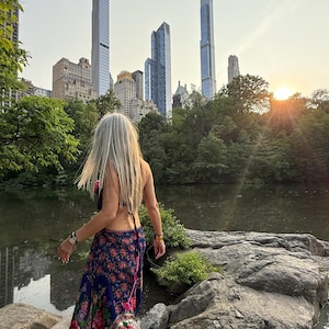 May include: A person with long gray hair stands near a pond in a park, with a city skyline in the background. They wear a floral print skirt and a small purse. The sun sets in the distance, casting a warm glow.