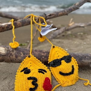 Puede incluir: Top de bikini de ganchillo amarillo con caras de emoji. Un top tiene gafas de sol, el otro tiene un corazón rojo y el número 3. El bikini se exhibe en una playa de arena con madera flotante.