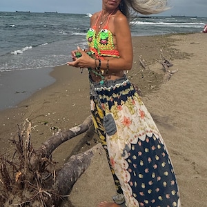May include: A colorful crochet top with a frog design, paired with a floral print skirt. The outfit is worn on a sandy beach with the ocean in the background. The top features orange, green, and yellow colors.